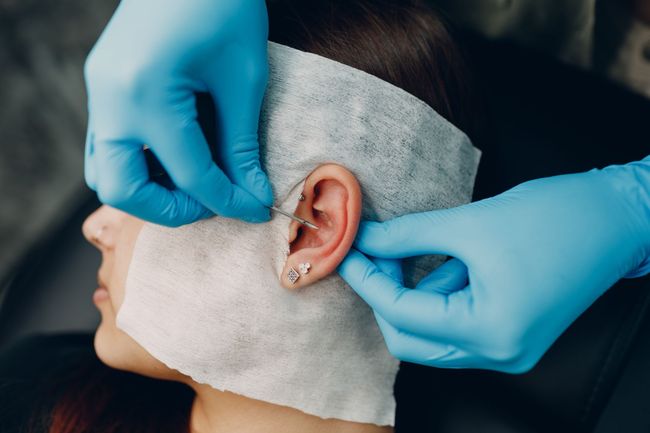 Gloved hands adjust an ear piercing on a person with a bandaged head.