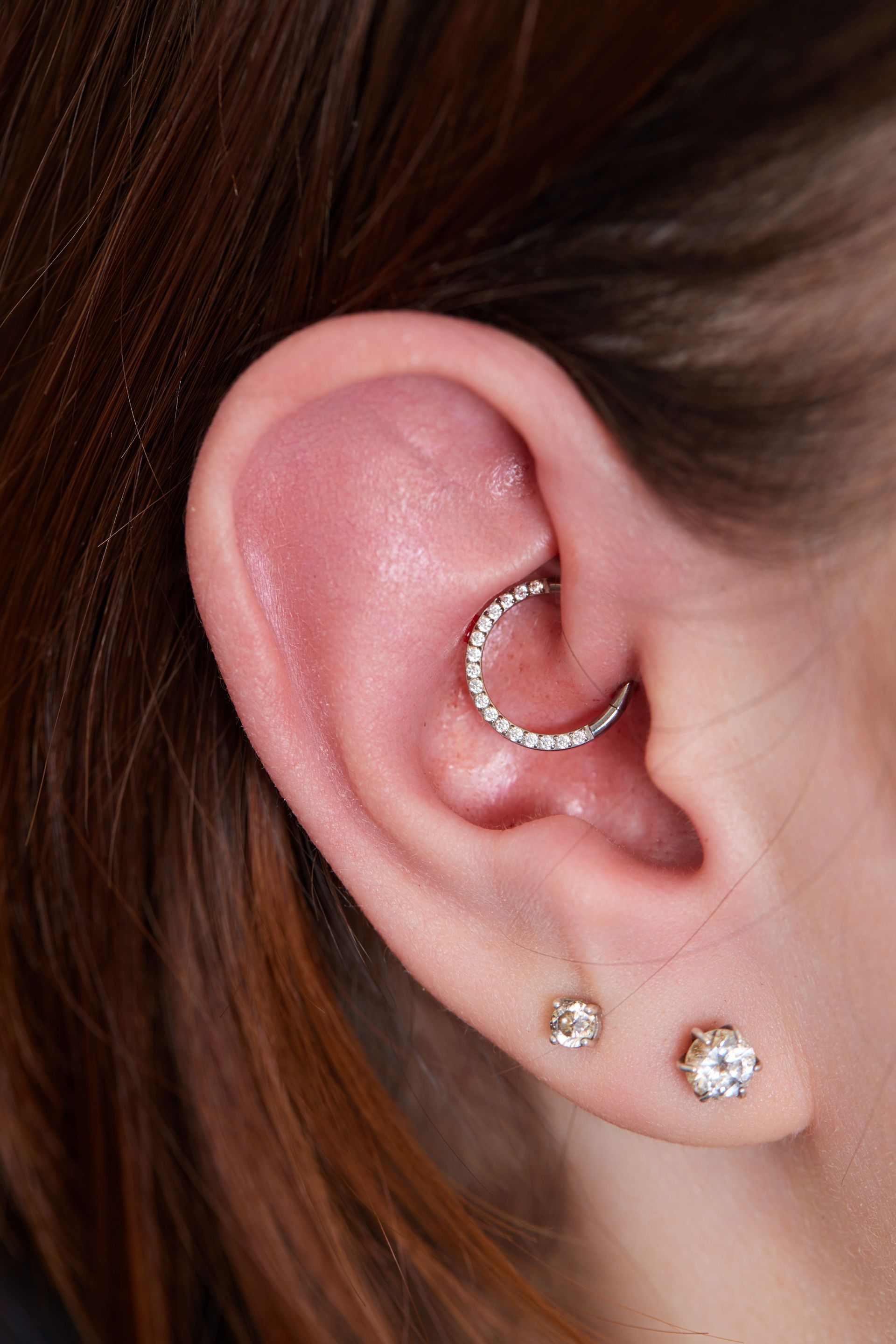 Close-up of an ear with a silver hoop and two stud earrings, against brown hair.