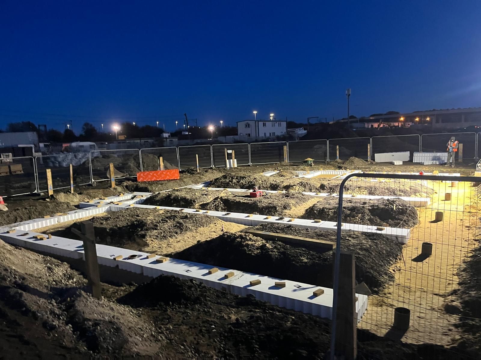 Nighttime construction site with lit foundation footings and excavated trenches under a dark blue sky