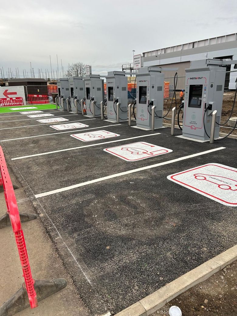 Outdoor EV charging stations with gray chargers on a paved lot, marked parking spaces, and a red tape barrier