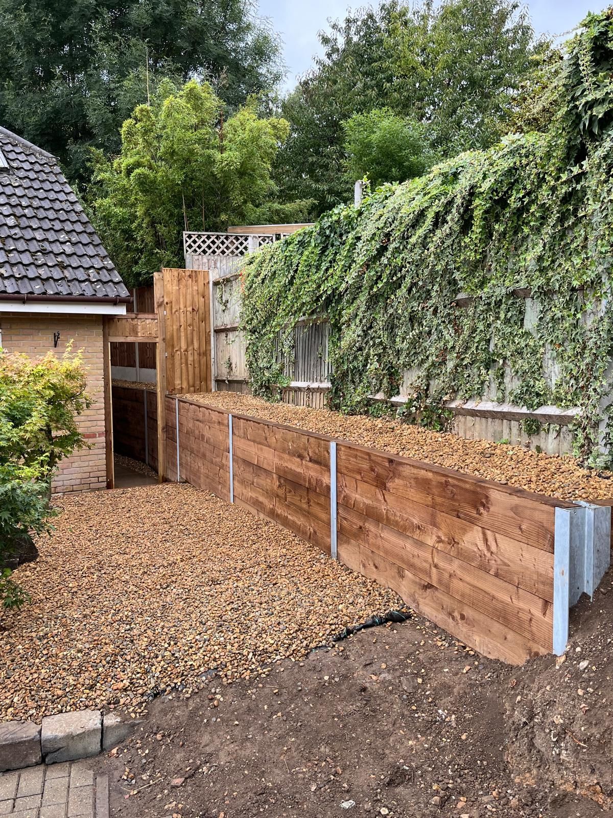 Backyard with a wooden raised bed, gravel path, and ivy-covered fence beside a house