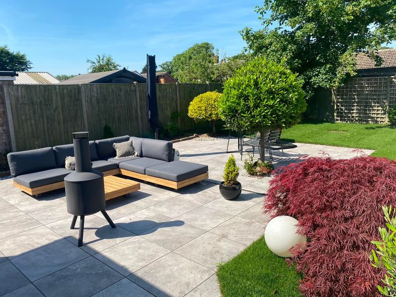 Modern backyard patio with gray outdoor seating, a fire pit, and red and green shrubs under a sunny sky