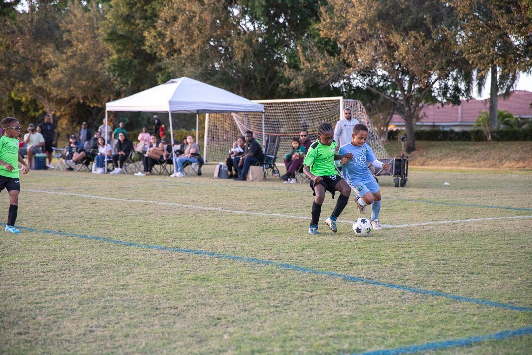 Youth soccer game with two players contesting the ball on a grassy field near spectators and a white tent.