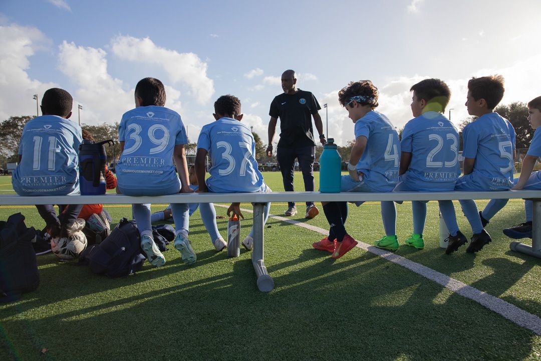 Youth soccer team in blue jerseys seated on a bench, listening to their coach on a sunny field.