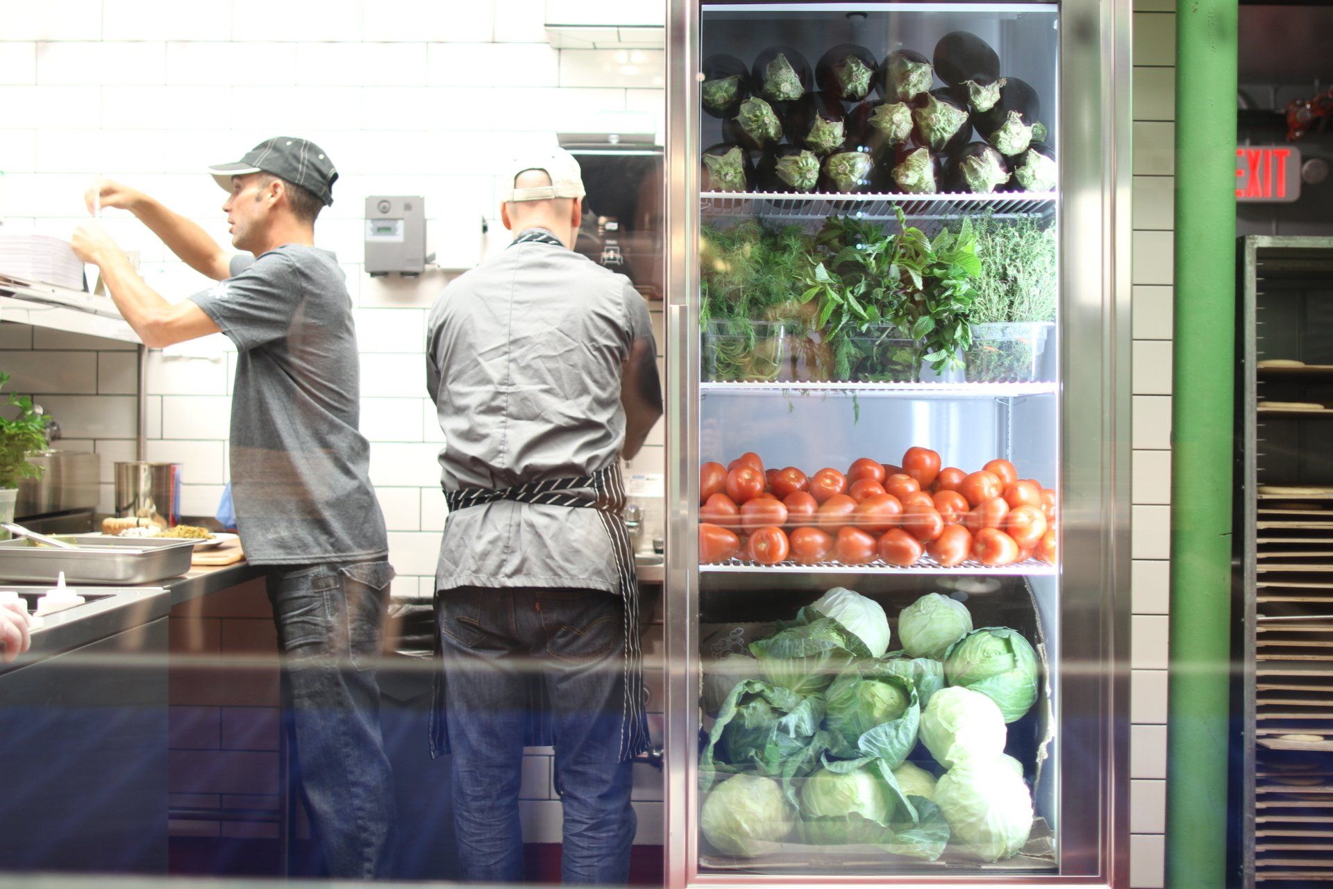 Two bonapita chefs working in a commercial kitchen with a refrigerator displaying produce: artichokes, tomatoes, cabbage, and greens.