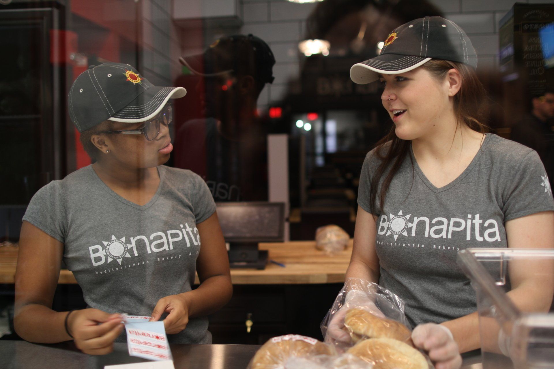 Two bonapita employees in gray shirts and caps behind a counter, one looking at a note, the other holding a bag.