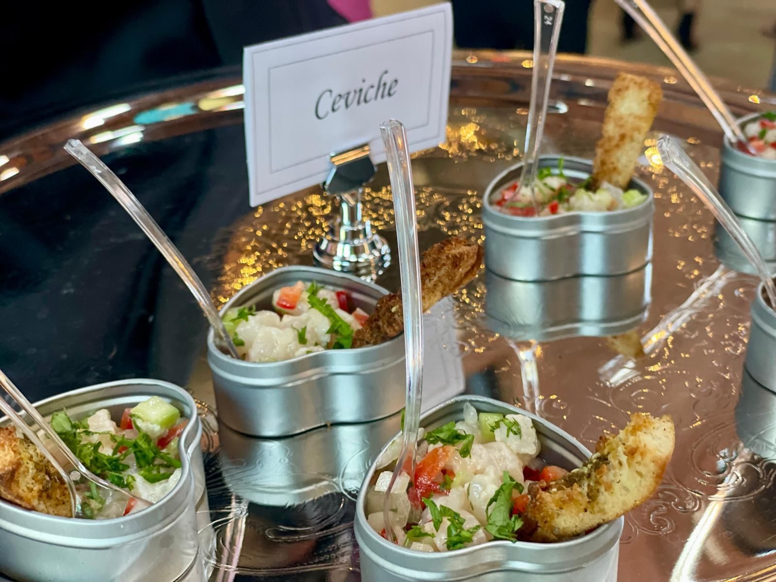 Ceviche in small metal containers on a silver platter, served with spoons and toasted bread.