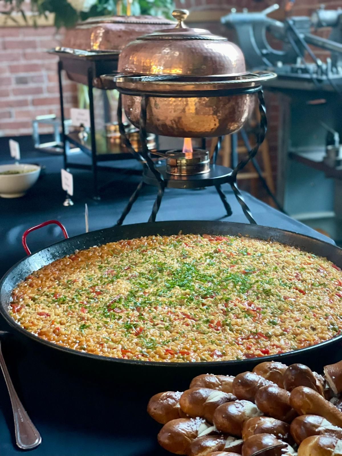 Buffet with a large paella pan, copper chafing dish, and rolls on a black tablecloth.