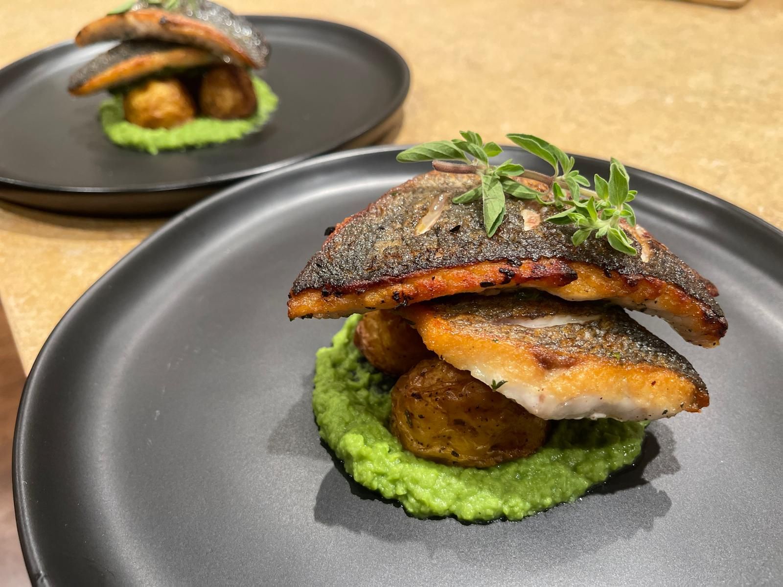 Two plated servings of pan-seared fish atop mashed peas, roasted potatoes, and herbs on black plates.