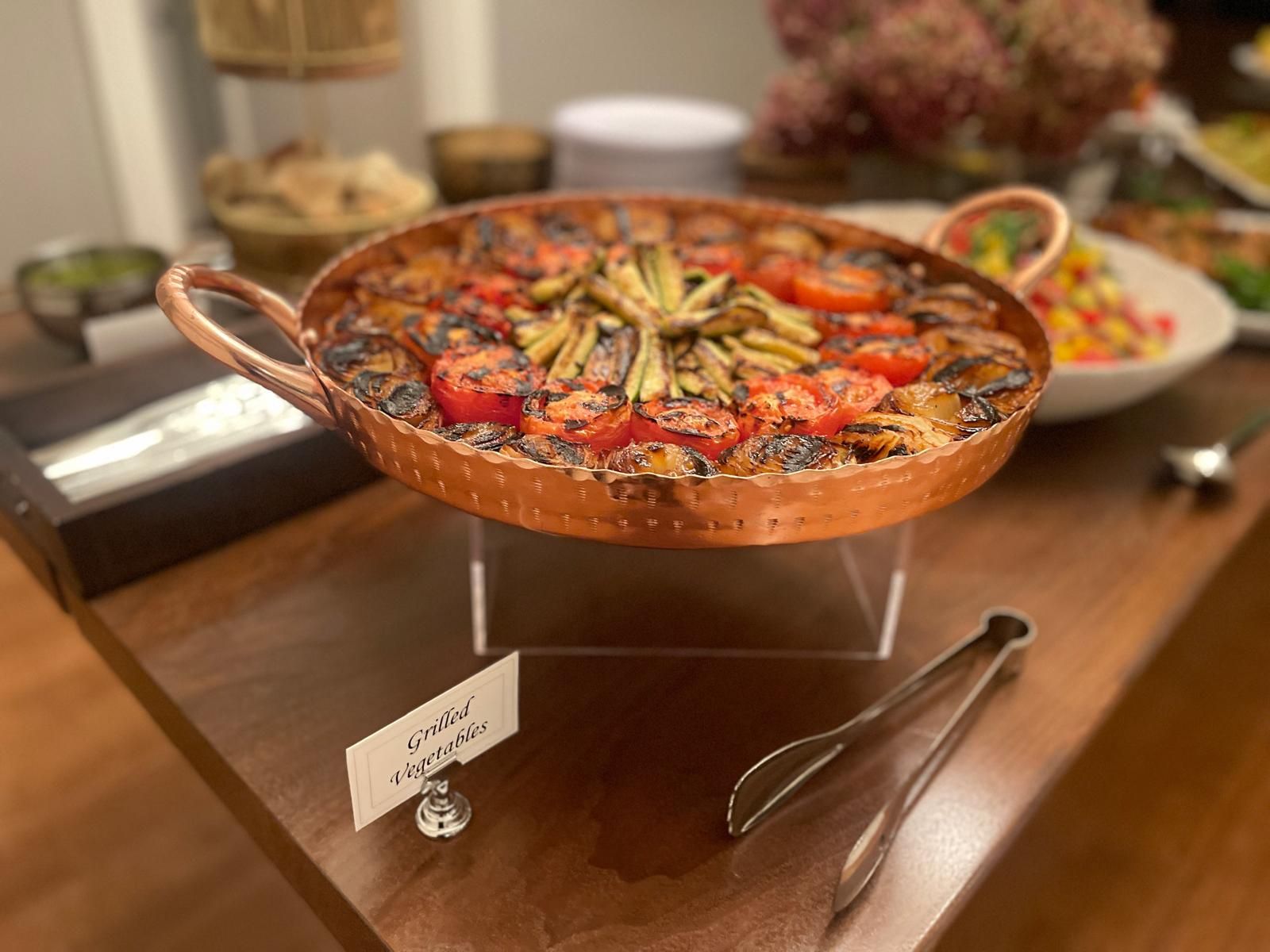 Copper pan of roasted vegetables on a buffet table, next to serving tongs and a label.