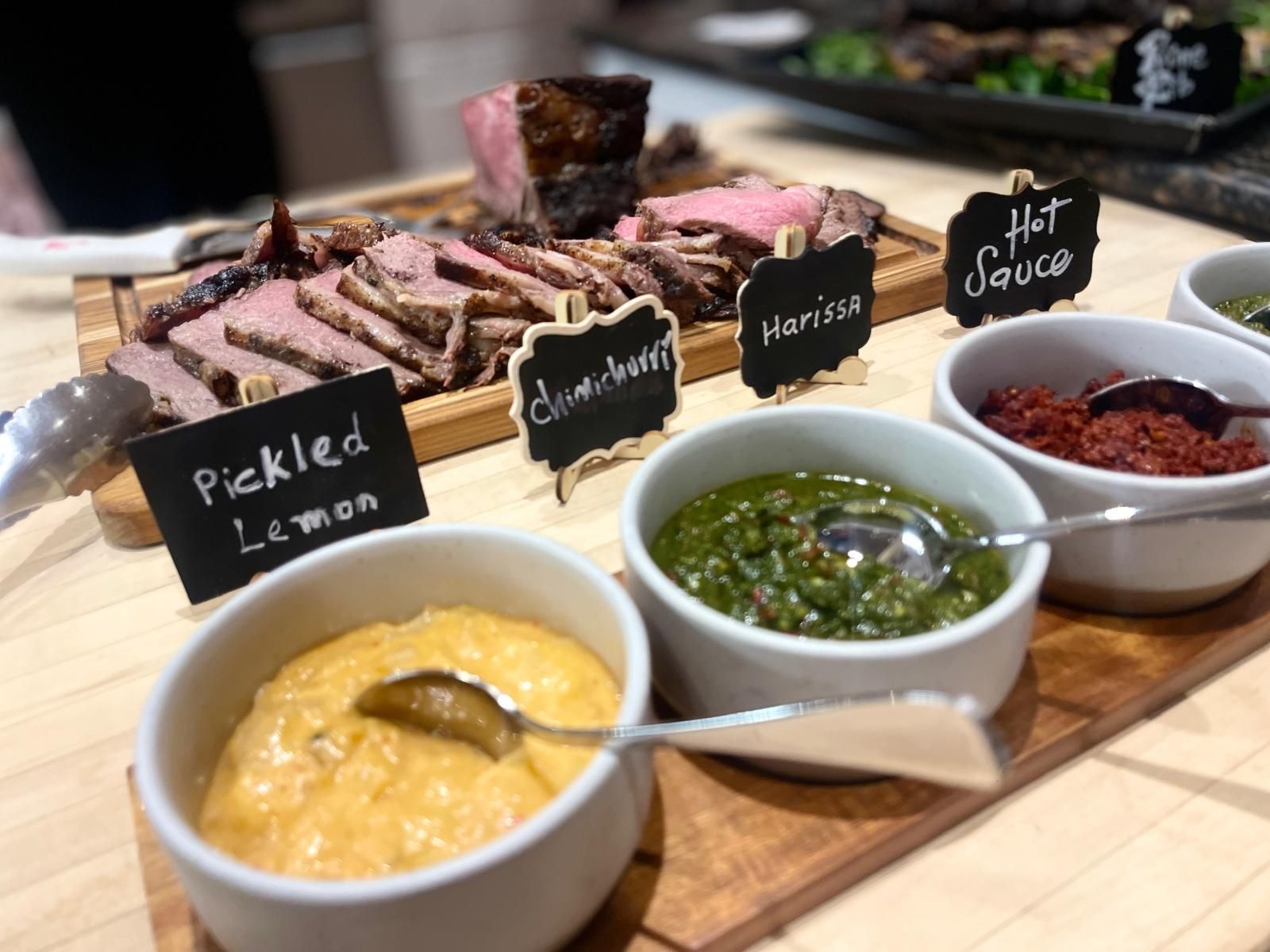 Sliced roast beef platter with small bowls of condiments: pickled lemon, chimichurri, harissa, and hot sauce.