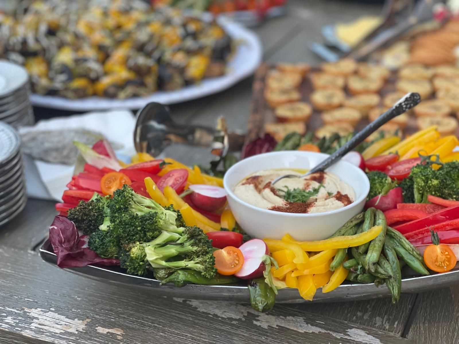Vegetable platter with hummus dip, surrounded by broccoli, peppers, and tomatoes.