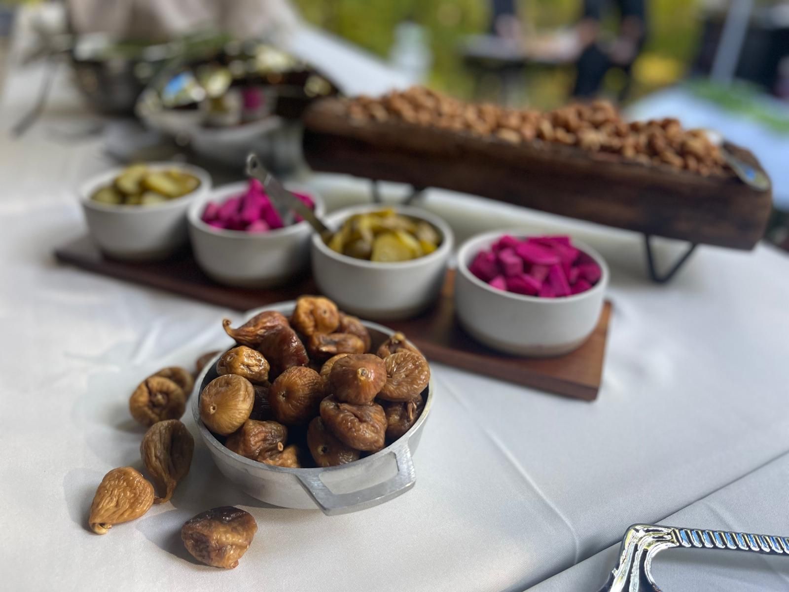 Table with food: nuts, pickles, and figs in white bowls and on a wooden tray, outdoors.