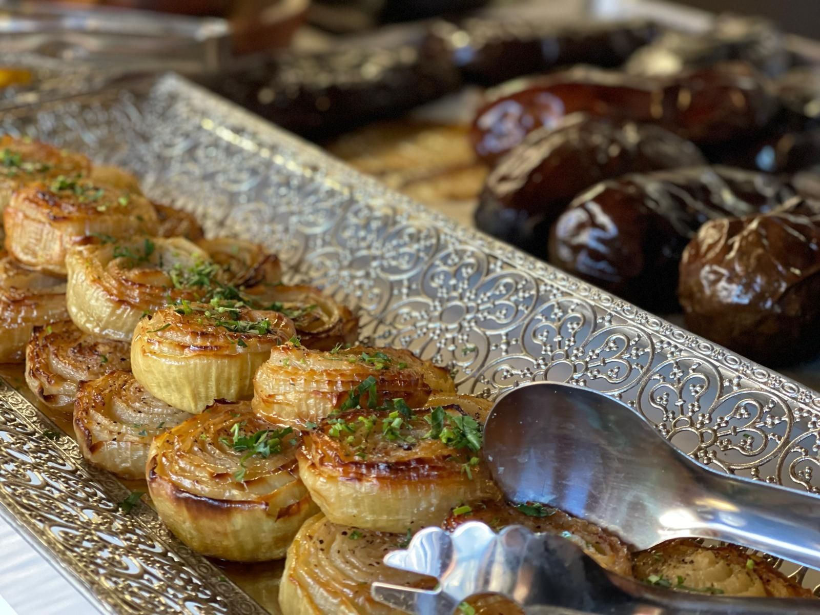 Roasted onions and eggplant on a decorative silver platter, tongs in foreground.