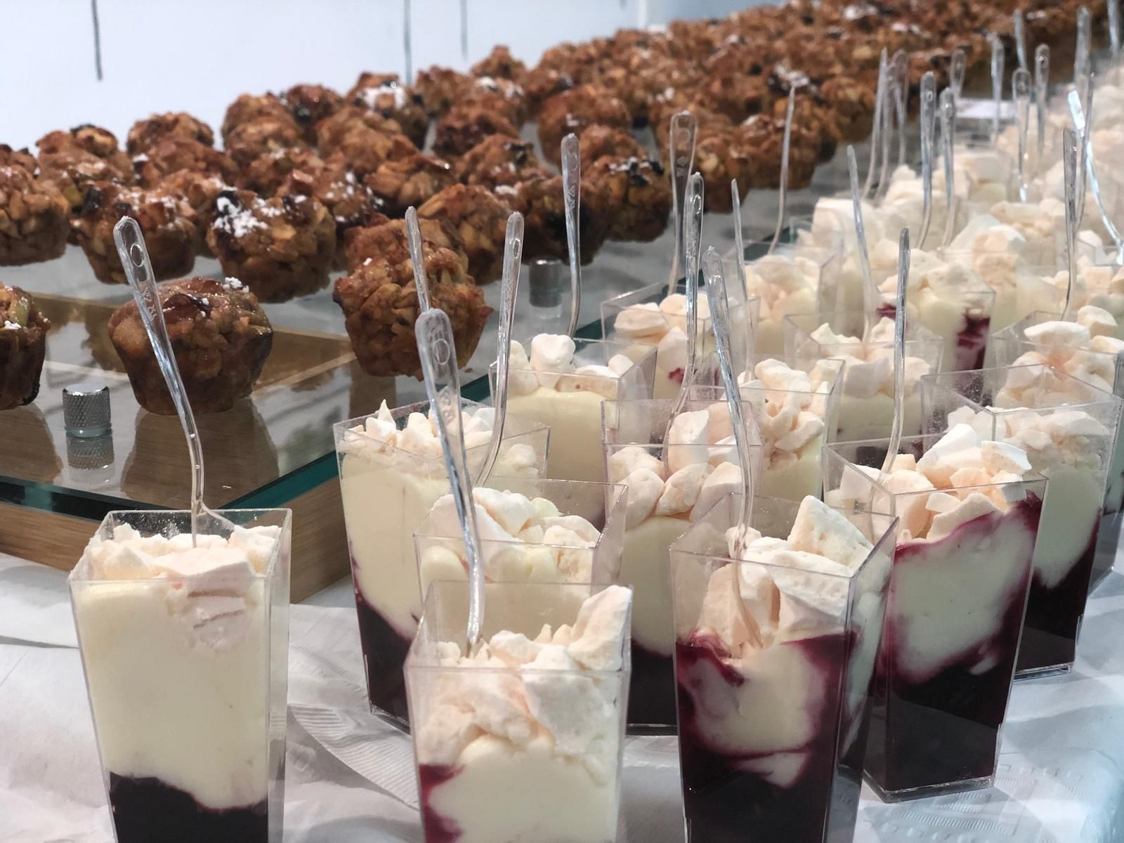 Dessert table with clear cups of layered cream and dark sauce, and muffins.