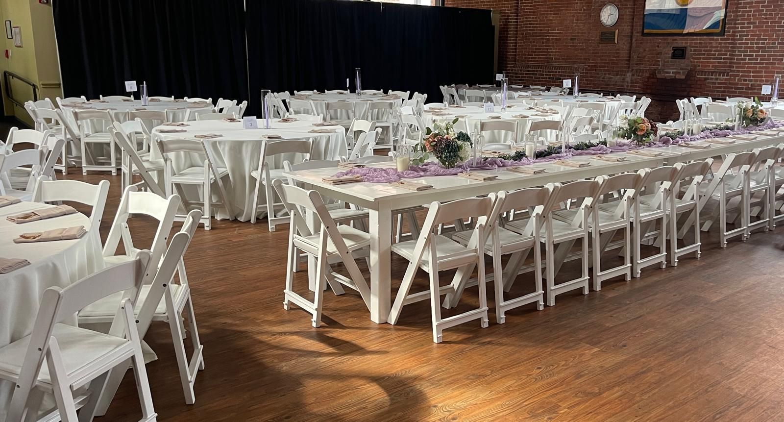 Event hall with white folding tables and chairs set for a gathering. Long table decorated with flowers.