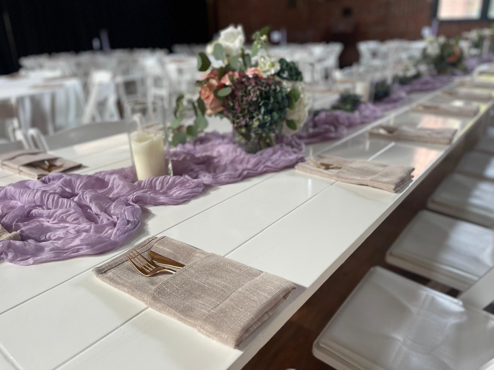 Long, white table set for a formal event with floral centerpieces, purple runner, and place settings.