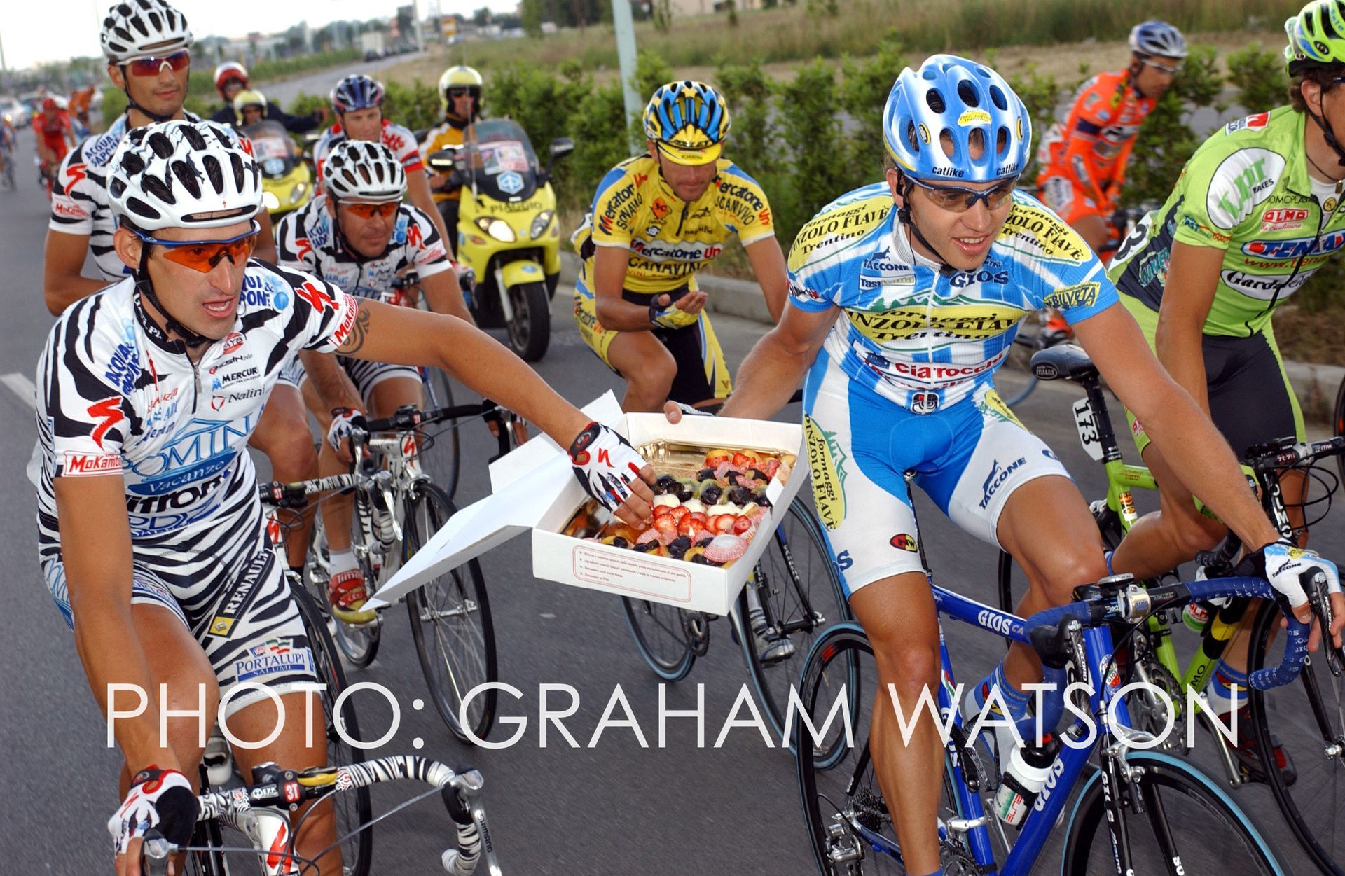 Cyclists racing closely together, one carrying a tray of cakes, in a crowded road race.