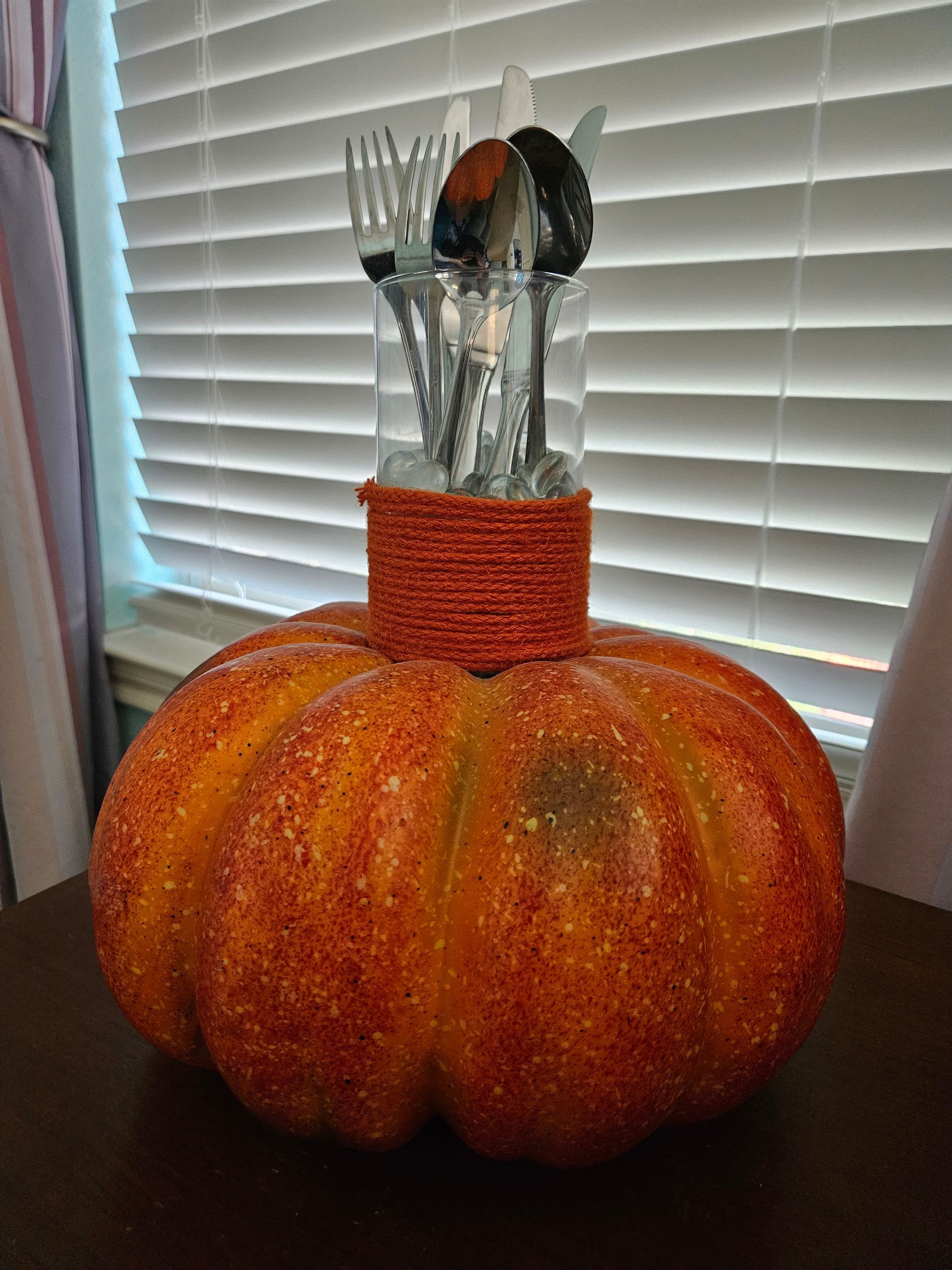 A pumpkin with utensils in it is sitting on a table in front of a window.