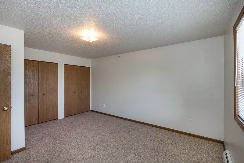 Empty beige bedroom with carpet, white walls, wooden closet doors, and a ceiling light.