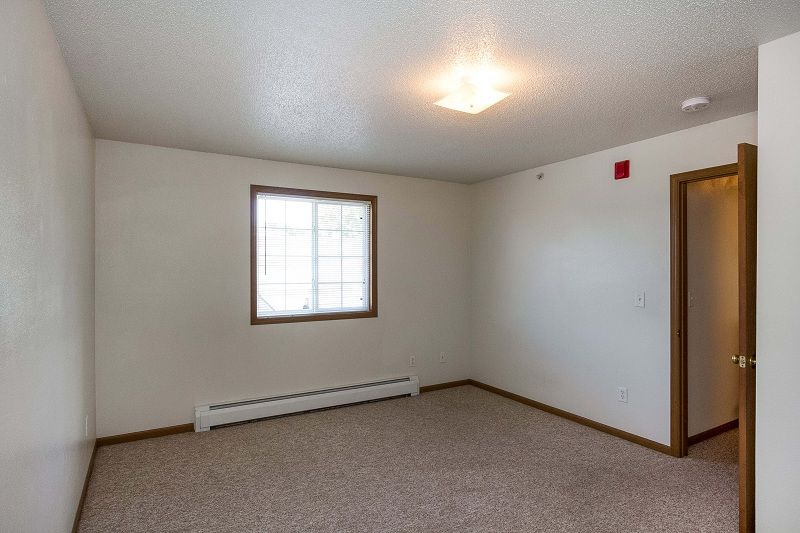 Empty beige bedroom with carpet, window, radiator, and open door to hallway