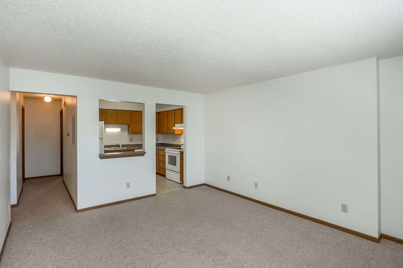 Empty beige living room with carpet, pass-through kitchen opening, and hallway entrance