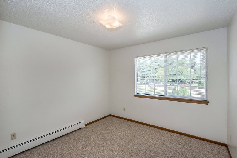Empty white bedroom with beige carpet, wood trim, and a window with blinds.