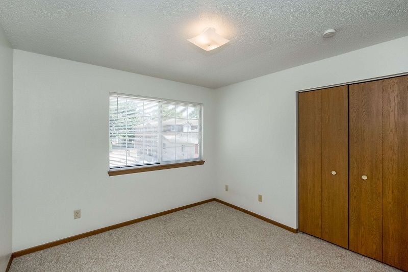 Empty carpeted bedroom with white walls, window blinds, and a wooden closet door.