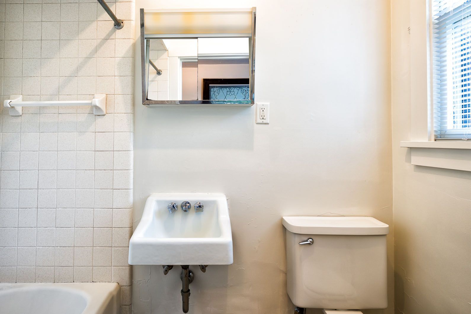 Small white bathroom with sink, toilet, mirror, tub edge, and window blinds.