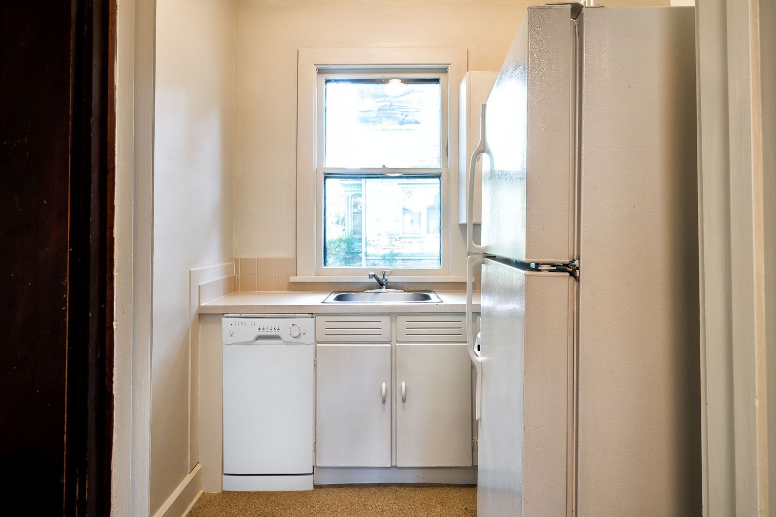 Narrow galley kitchen with white cabinets, sink under a window, and a white refrigerator on the right.