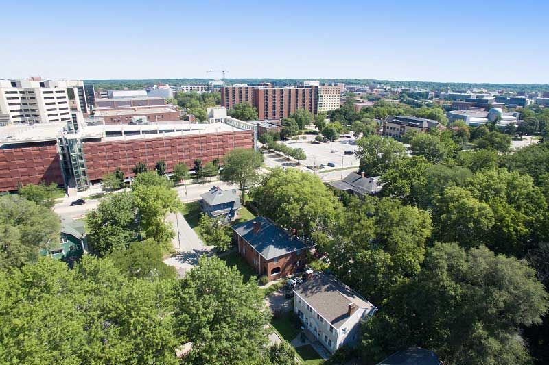 Aerial view of a tree-lined urban campus with brick buildings and a distant city skyline