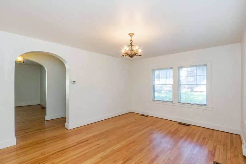 Empty living room with hardwood floors, white walls, arched doorway, window, and ceiling light fixture