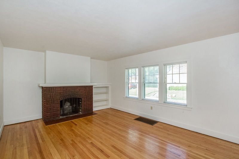 Empty living room with brick fireplace, hardwood floors, and large windows