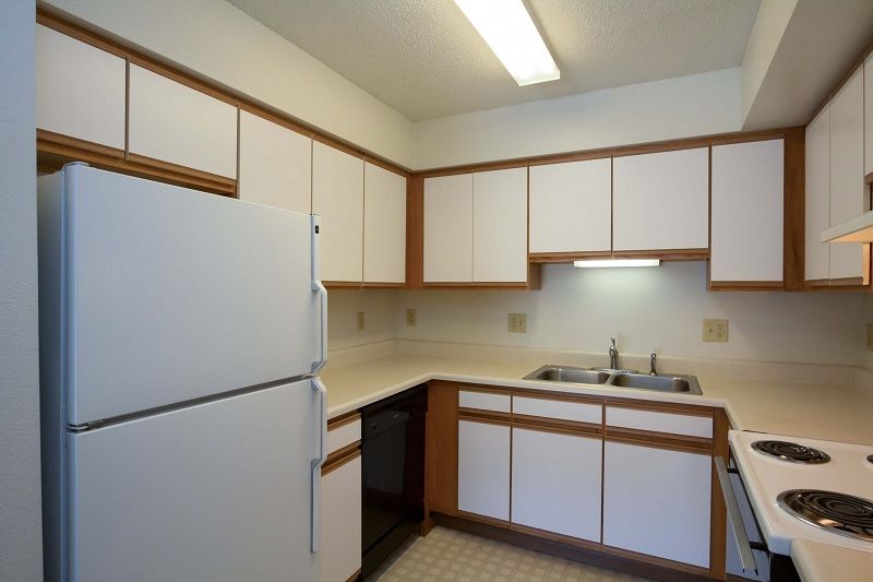 Small kitchen with white cabinets, refrigerator, stove, sink, and beige countertops under fluorescent lighting.