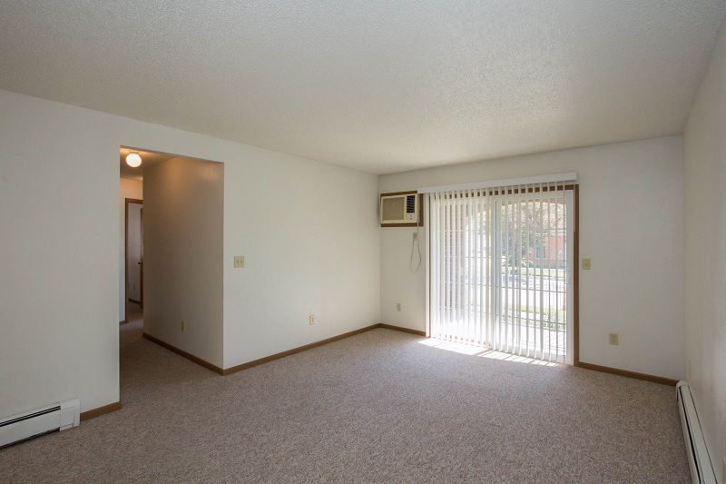 Empty carpeted living room with sliding glass door, window AC unit, and doorway to a hall