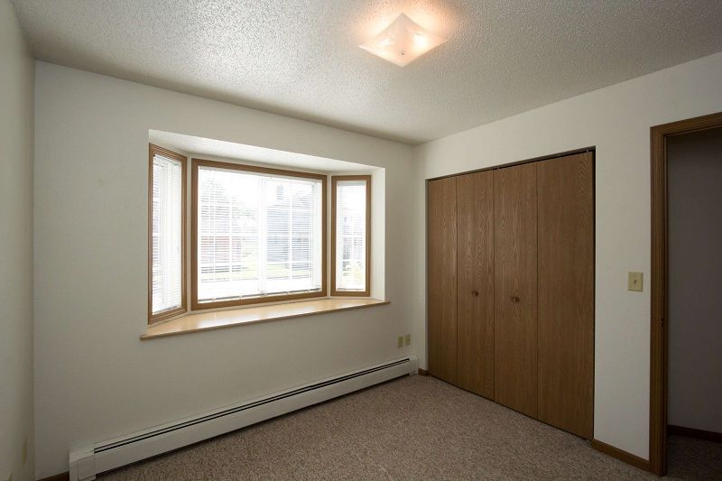 Empty bedroom with beige carpet, white walls, a window, and wooden closet doors.