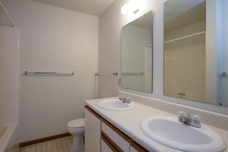 Small bathroom with double sinks, mirrors, toilet, and towel bar in a beige room.