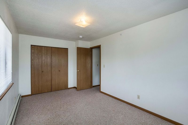 Empty beige bedroom with white walls, carpet, closet doors, and a window on the left.