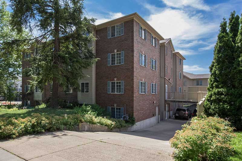 Brick apartment building with landscaped entrance, driveway, and parked car on a sunny day