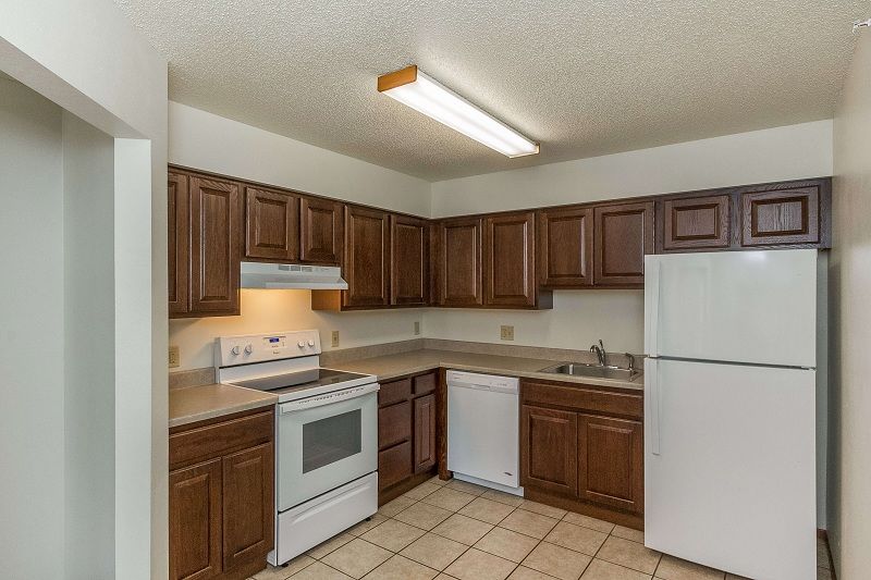 Empty kitchen with brown cabinets, white appliances, tan tile floor, and fluorescent ceiling light