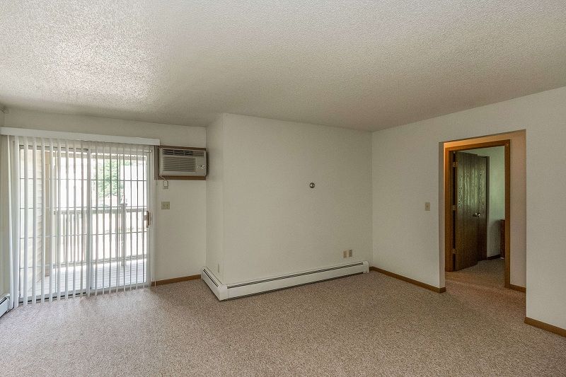 Empty beige living room with carpet, sliding glass door, wall AC, and open doorway to hallway
