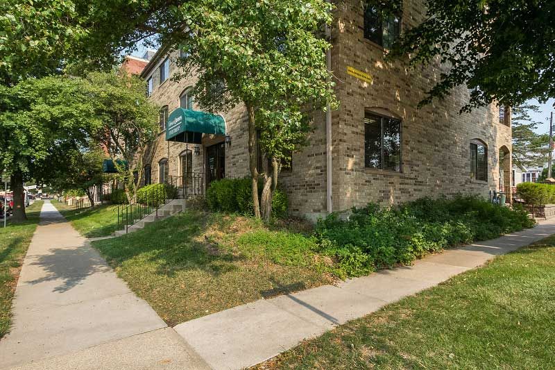 Brick apartment building with green awning beside a tree-lined sidewalk and grassy corner