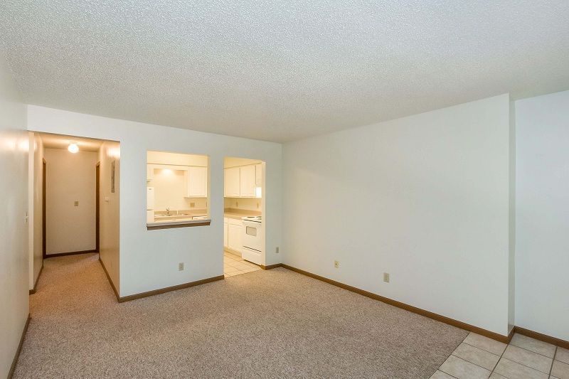 Empty beige apartment living room with carpet, pass-through opening to kitchen, and tiled entryway