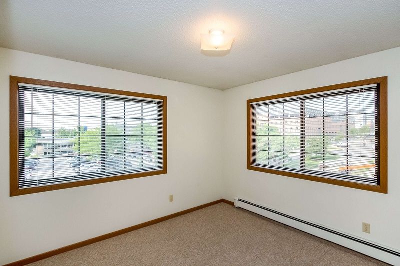 Empty carpeted room with two windows, wood blinds, and beige walls.
