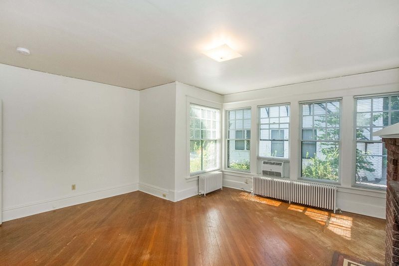 Empty living room with hardwood floors, white walls, large bay windows, and a radiator under the windows.