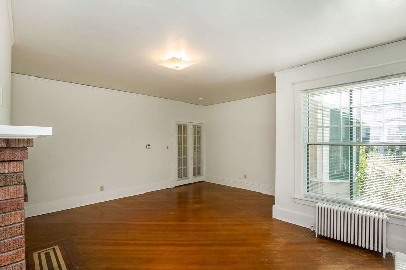 Empty living room with hardwood floors, white walls, ceiling light, and a large window with radiator.