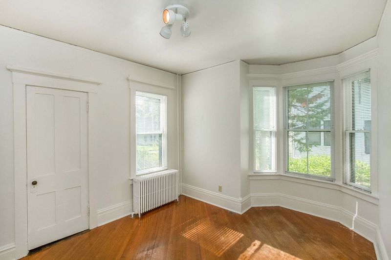 Empty white room with hardwood floors, bay windows, and a radiator near the left wall.