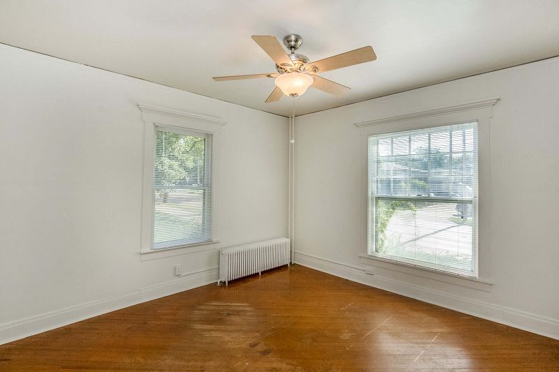 Empty room with hardwood floor, white walls, ceiling fan, and two windows with blinds.