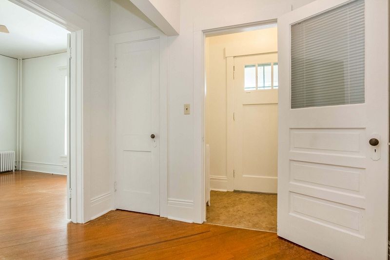 Bright empty hallway with white doors and wood floors, one door open to a beige room.