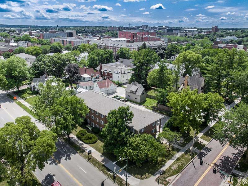 Aerial view of a leafy neighborhood with houses, streets, and a distant city skyline under a partly cloudy sky